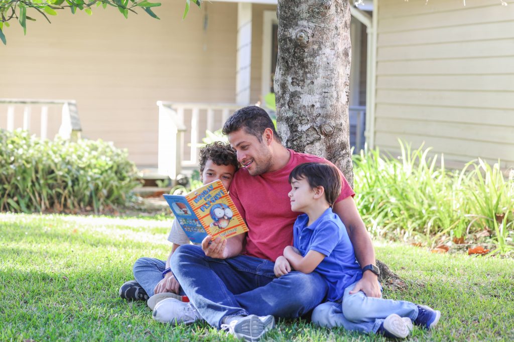 New Worlds Reading Families-149 A father sits beneath a tree with his two sons cuddled next to him. The father reads a book aloud to the children; one child smiles, while one child looks focused, sticking his tongue out while he thinks